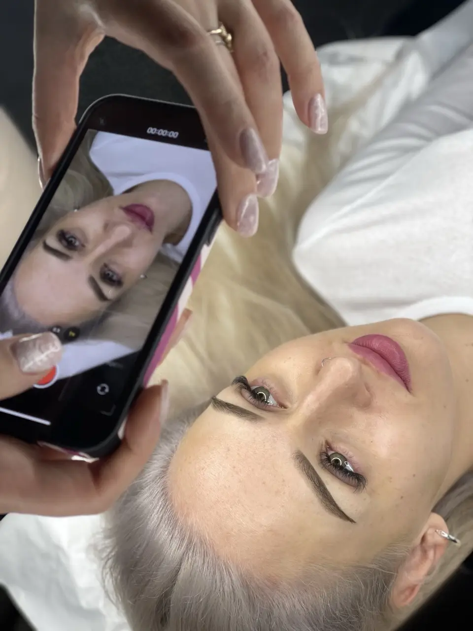 projektowanie stron www Kobieta z platynowo-blond włosami leży twarzą do góry, a fotograf w Augustowie uwiecznia jej wizerunek. Smartfon nad nią pokazuje jej odbicie, podkreślając jej lekki makijaż i biały top. fotografia filmowanie augustów