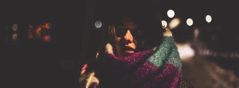 projektowanie stron www Osoba stoi w ciemności, ma na sobie ciemny kapelusz i wielokolorowy szalik owinięty wokół twarzy. Rozmyte światła w tle przywołują sceny z twórczości filmowca, tworząc tajemniczą atmosferę przypominającą augustowskie noce. fotografia filmowanie augustów