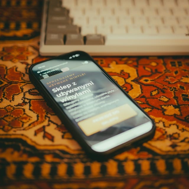 projektowanie stron www Smartfon wyświetlający stronę internetową w języku polskim spoczywa na wzorzystym dywanie. W tle częściowo widoczna jest klawiatura. Scena ma ciepły ton kolorów. fotografia filmowanie augustów