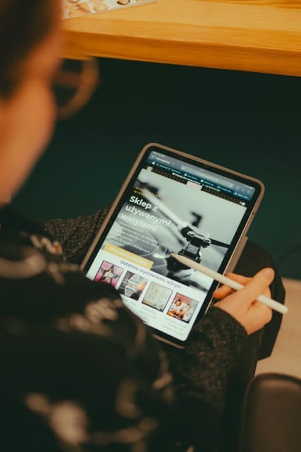 projektowanie stron www Osoba trzymająca tablet z rysikiem. Na ekranie wyświetlana jest strona internetowa z tekstem w języku polskim i obrazami związanymi ze sklepem. Osoba ma na sobie wzorzysty sweter i okulary. W tle widoczna jest drewniana krawędź stołu. fotografia filmowanie augustów