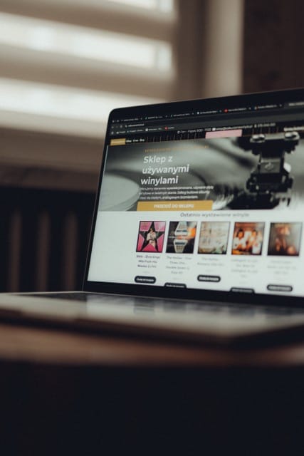 projektowanie stron www Laptop wyświetlający stronę internetową sklepu z płytami winylowymi w języku polskim. Na ekranie wyświetlane są obrazy płyt winylowych i pokrewnych kategorii. Sceneria wydaje się być w pomieszczeniu, a światło wpada przez żaluzje w tle. fotografia filmowanie augustów