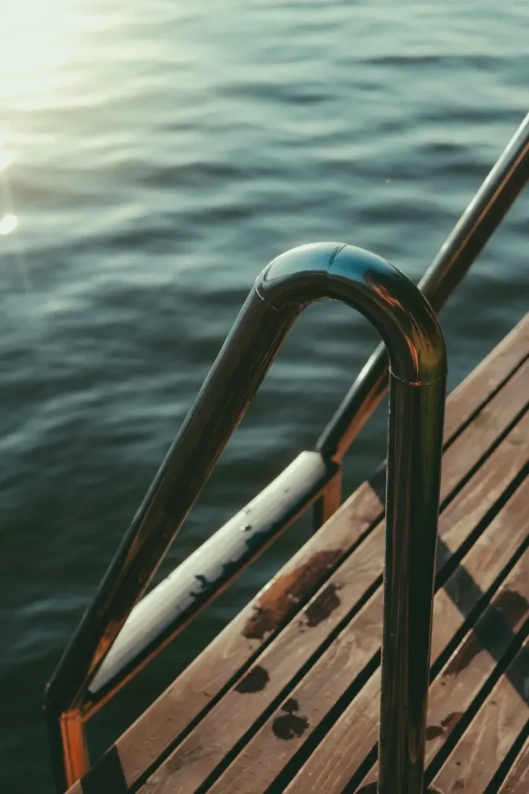 projektowanie stron www Metalowa poręcz i drewniane stopnie prowadzą do spokojnego zbiornika wodnego, lśniącego w świetle słonecznym. Odblaskowe powierzchnie podkreślają spokojne otoczenie, przypominające ujęcie fotografa Augustowa. Sugeruje spokojne miejsce do pływania lub relaksu nad brzegiem wody. fotografia filmowanie augustów