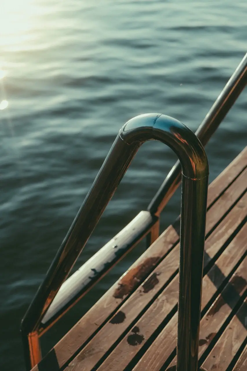 projektowanie stron www Metalowa poręcz i drewniane stopnie prowadzą do spokojnego zbiornika wodnego, lśniącego w świetle słonecznym. Odblaskowe powierzchnie podkreślają spokojne otoczenie, przypominające ujęcie fotografa Augustowa. Sugeruje spokojne miejsce do pływania lub relaksu nad brzegiem wody. fotografia filmowanie augustów