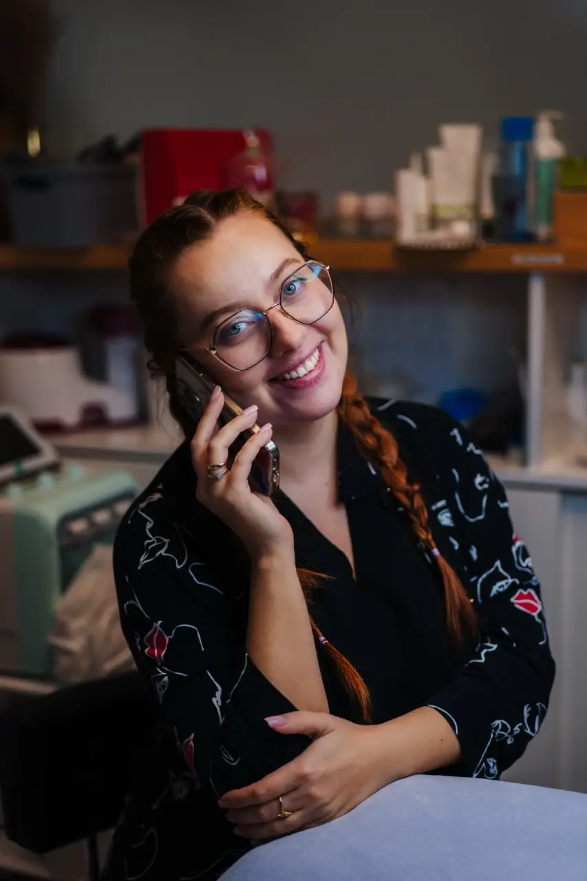 projektowanie stron www Kobieta w okularach i z warkoczami uśmiecha się, rozmawiając przez smartfon. Ma na sobie wzorzysty czarny top i opiera się o stół. W tle znajdują się półki z różnymi butelkami i pojemnikami. fotografia filmowanie augustów