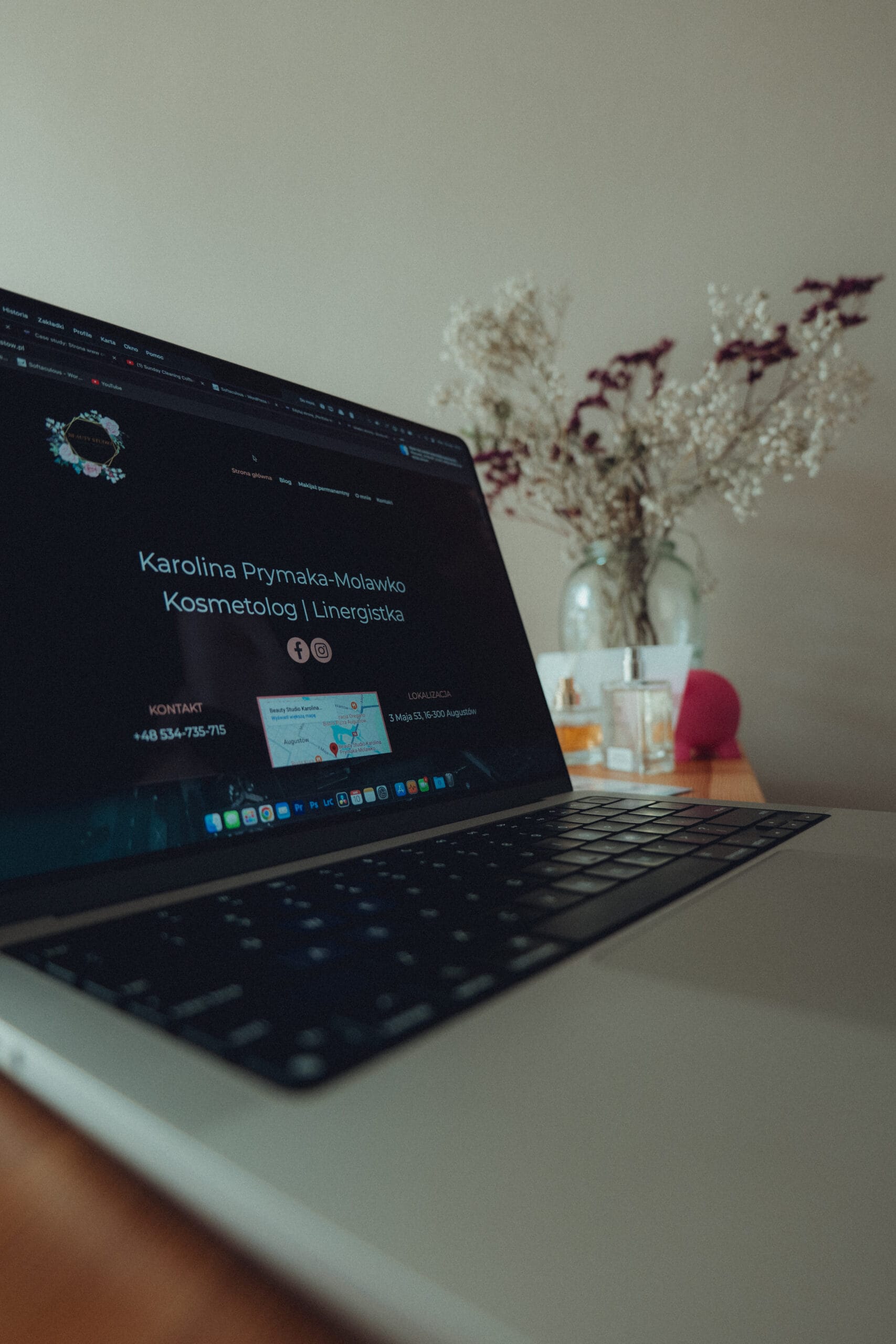 projektowanie stron www Na laptopie wyświetla się strona internetowa z danymi kontaktowymi „Karolina Prymaka-Molakwo Kosmetolog i Linergistka”. W tle na drewnianym stole znajduje się rozmazany wazon z suszonymi kwiatami. fotografia filmowanie augustów