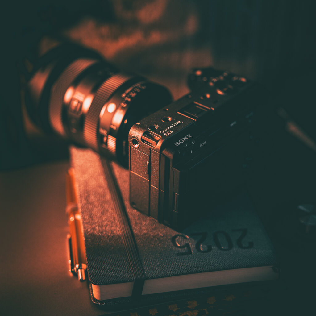 projektowanie stron www Aparat Sony z dużym obiektywem leży na zamkniętym planerze na rok 2024. Obok planera leży długopis. Scena jest ciepło oświetlona, co tworzy przytulną i kreatywną atmosferę. fotografia filmowanie augustów