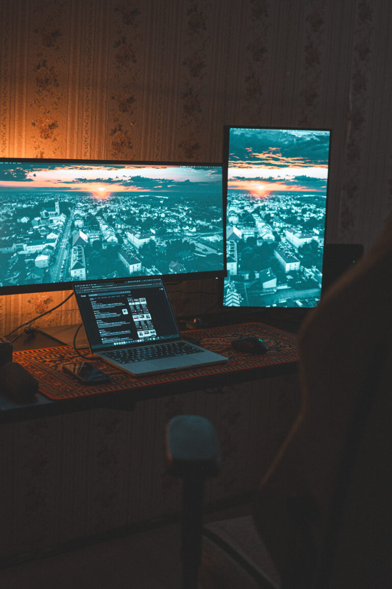 projektowanie stron www Domowe biuro z dwoma monitorami, jednym poziomym i jednym pionowym, oba wyświetlające obraz krajobrazowy. Laptop jest otwarty na biurku między nimi, wyświetlając program graficzny. Pokój jest słabo oświetlony ciepłym blaskiem lampy. fotografia filmowanie augustów