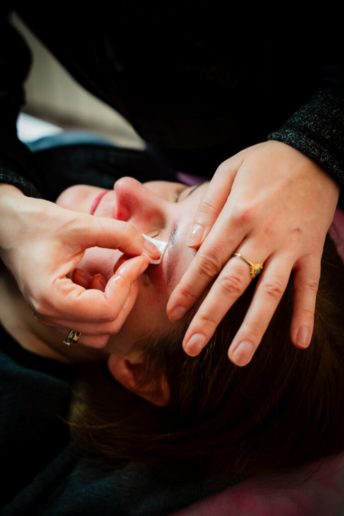 projektowanie stron www Osoba kładzie się, podczas gdy inna osoba delikatnie usuwa pasek wosku z okolic brwi, kształtując brwi. Zbliżenie pokazuje ostrożne ruchy dłoni i skupienie na brwi. fotografia filmowanie augustów