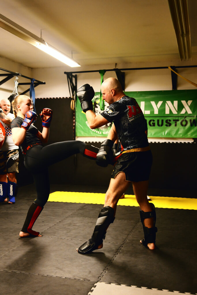 projektowanie stron www Dwie osoby w strojach ochronnych walczą na siłowni sztuk walki, jedna osoba wykonuje kopnięcie z przodu, a druga podnosi kolano, aby zablokować. W tle widać innych i zielony baner treningowy. fotografia filmowanie augustów