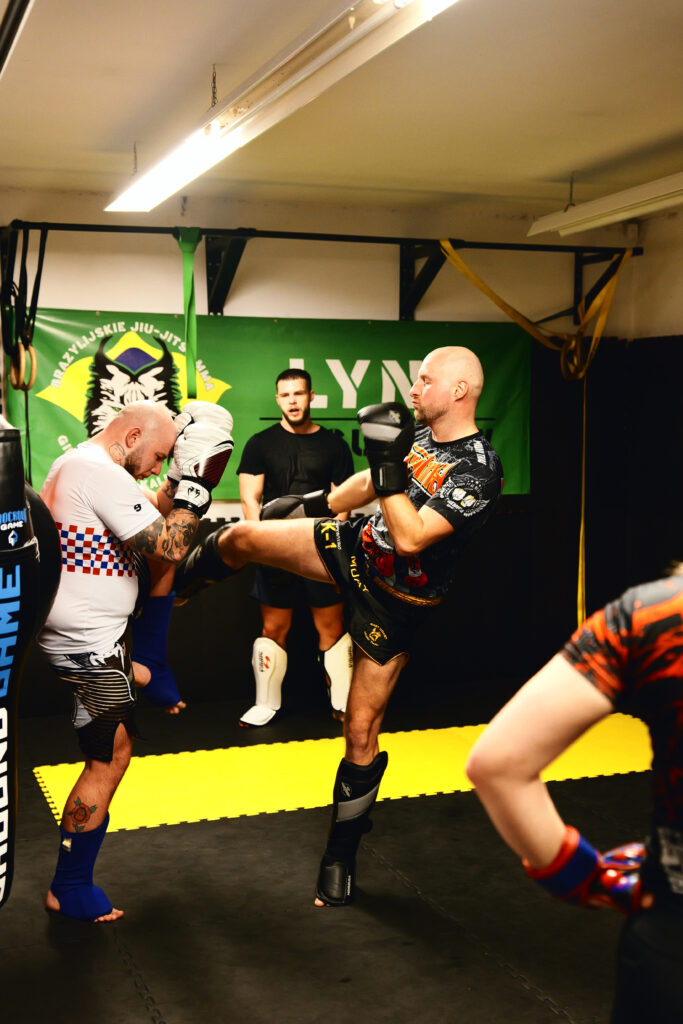 projektowanie stron www Grupa mężczyzn ćwiczy kickboxing na siłowni. Jeden z nich jest w trakcie kopnięcia, celując w przeciwnika, który blokuje go ramionami. Inni, w rękawicach i ochraniaczach, obserwują lub przygotowują się w pobliżu. Na ścianie wisi zielony baner siłowni. fotografia filmowanie augustów