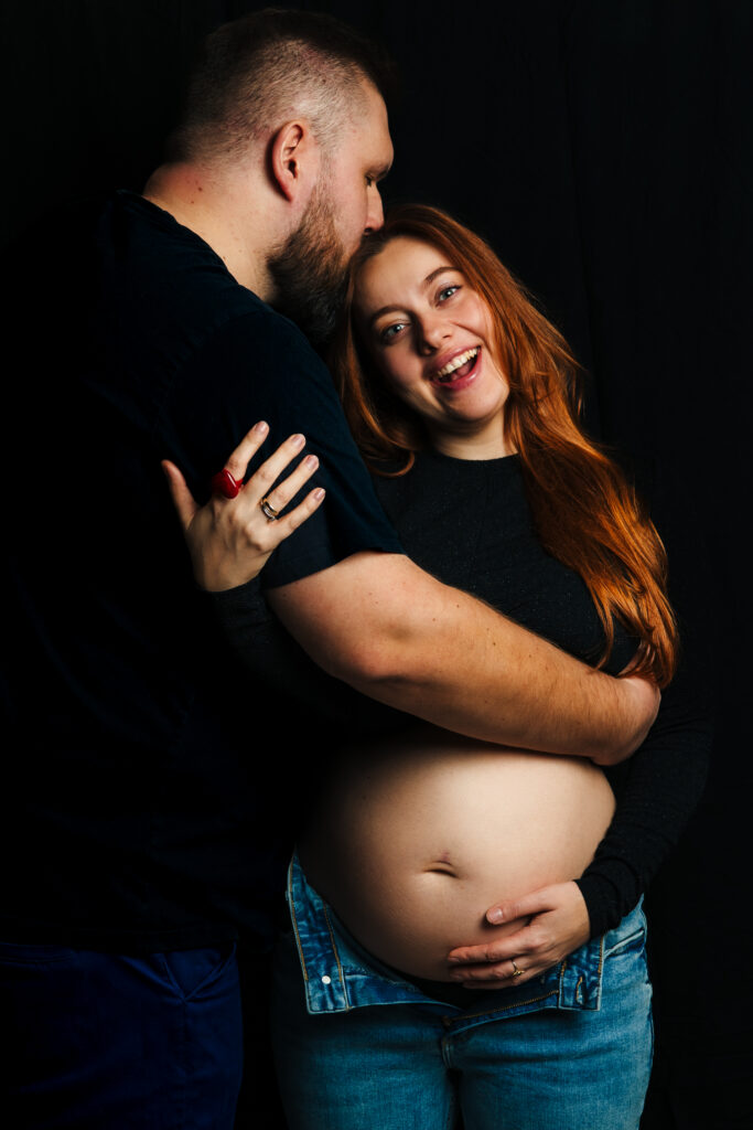 projektowanie stron www Mężczyzna obejmuje i całuje uśmiechniętą ciężarną kobietę w czoło. Kobieta trzyma się za nagi brzuch jedną ręką, ma na sobie dżinsy i ciemną koszulę z długim rękawem, stoi na czarnym tle. fotografia filmowanie augustów
