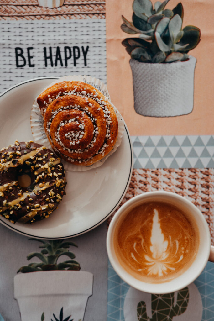 projektowanie stron www Filiżanka kawy z latte art położona obok pączka oblanego czekoladą i bułeczki cynamonowej na talerzu, na wzorzystym obrusie z sukulentami i napisem BE HAPPY. fotografia filmowanie augustów