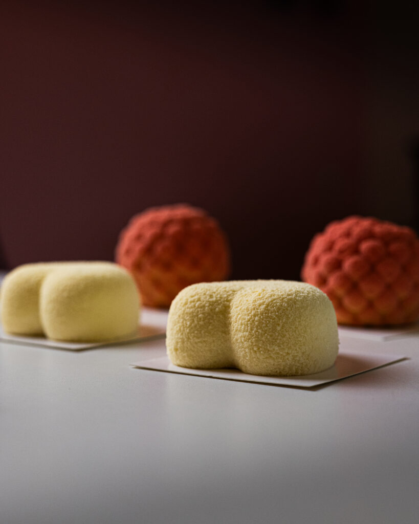 projektowanie stron www Dwa żółte, okrągłe desery musowe o aksamitnej konsystencji umieszczono na białych kartach na pierwszym planie. W tle dwa teksturowane, różowo-pomarańczowe desery przypominające szyszki sosnowe leżą na jasnej powierzchni. fotografia filmowanie augustów