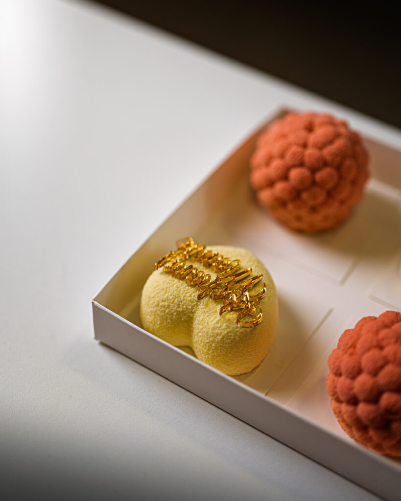 projektowanie stron www Zbliżenie białego pudełka zawierającego trzy eleganckie ciastka, w tym żółty deser w kształcie serca ze złotym napisem „Wszystkiego najlepszego z okazji urodzin” oraz dwa okrągłe, różowe ciastka o teksturowanej fakturze. fotografia filmowanie augustów